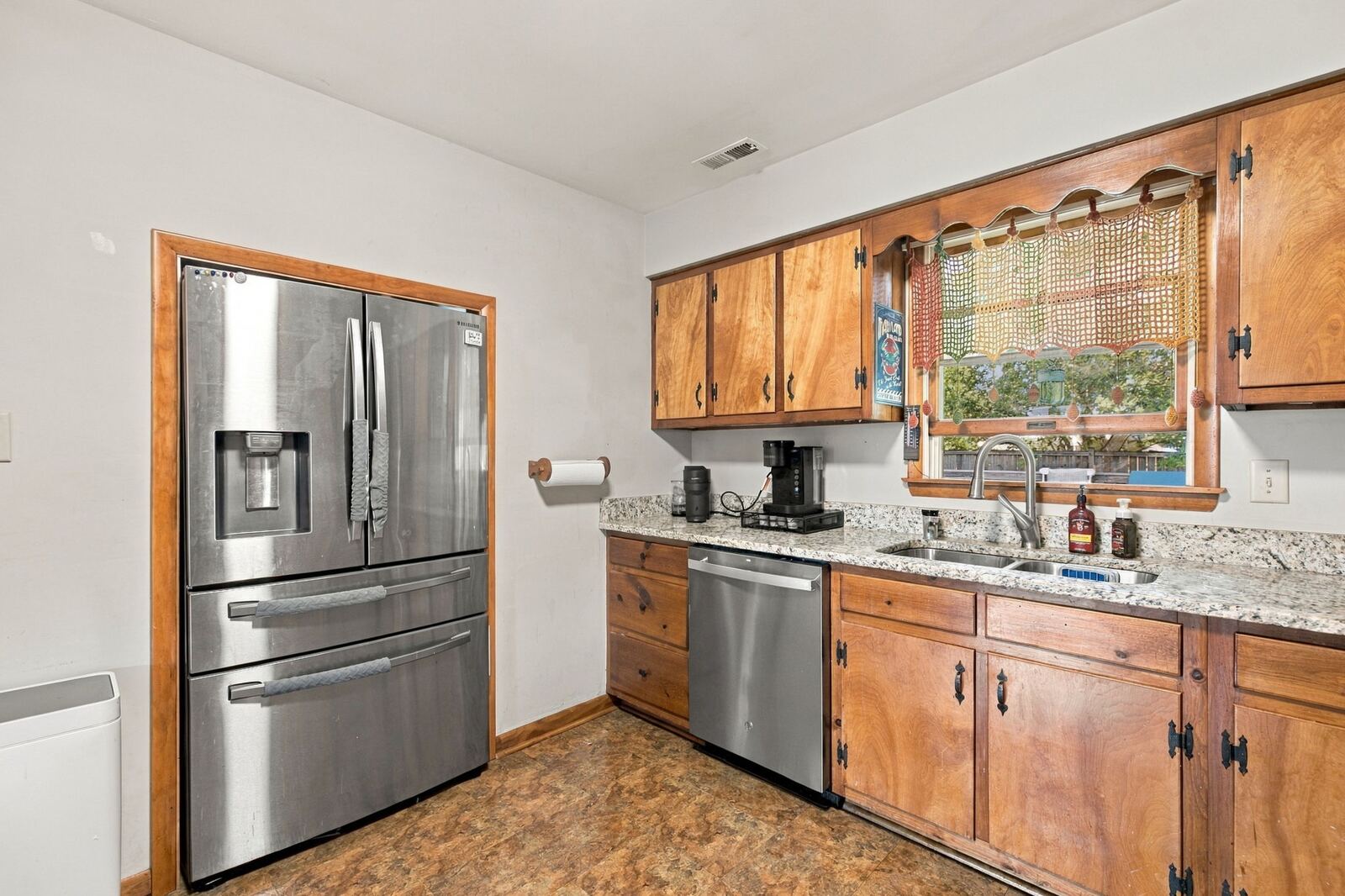 Kitchen sink below a window, framed by wood cabinets, with a stainless refrigerator and dishwasher.