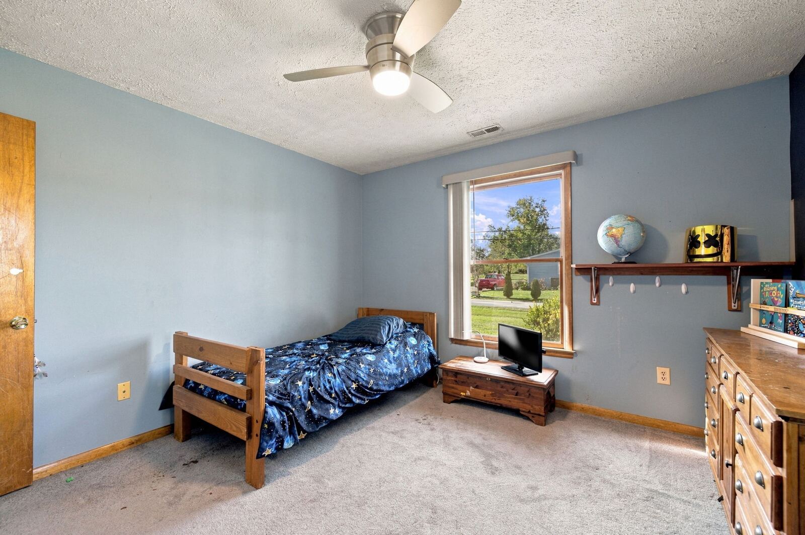 Bedroom with light blue walls, a ceiling fan, a twin bed, carpet and a window with a wall shelf.