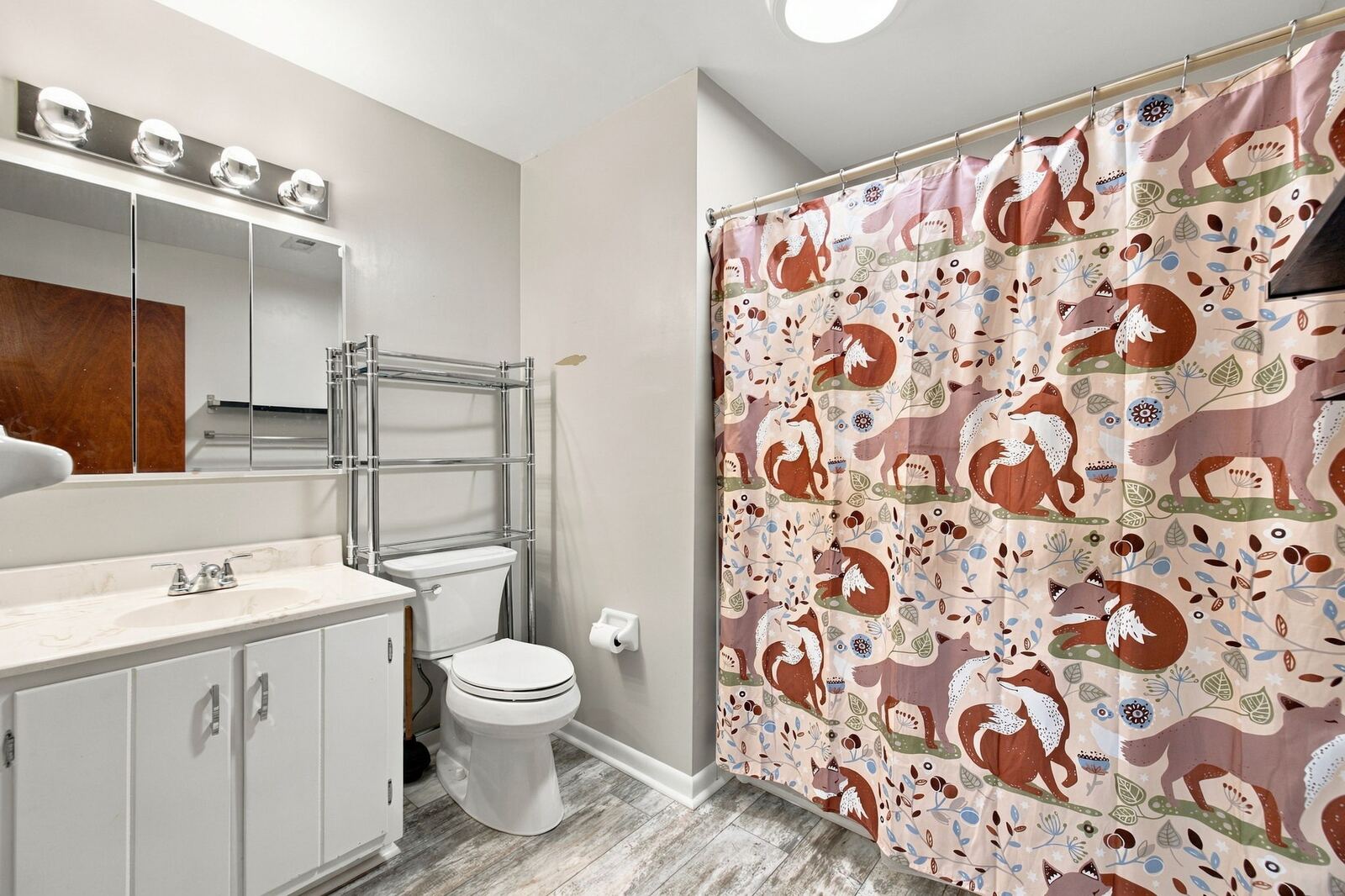 Hall bath with a white vanity, a lighted mirror, a toilet, a shower curtain and wire shelving.