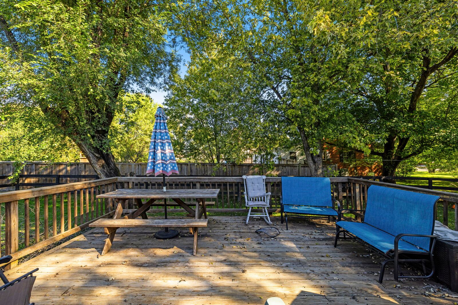 Wood rear deck with railings, a picnic table and patio chairs, with mature shade trees beyond.